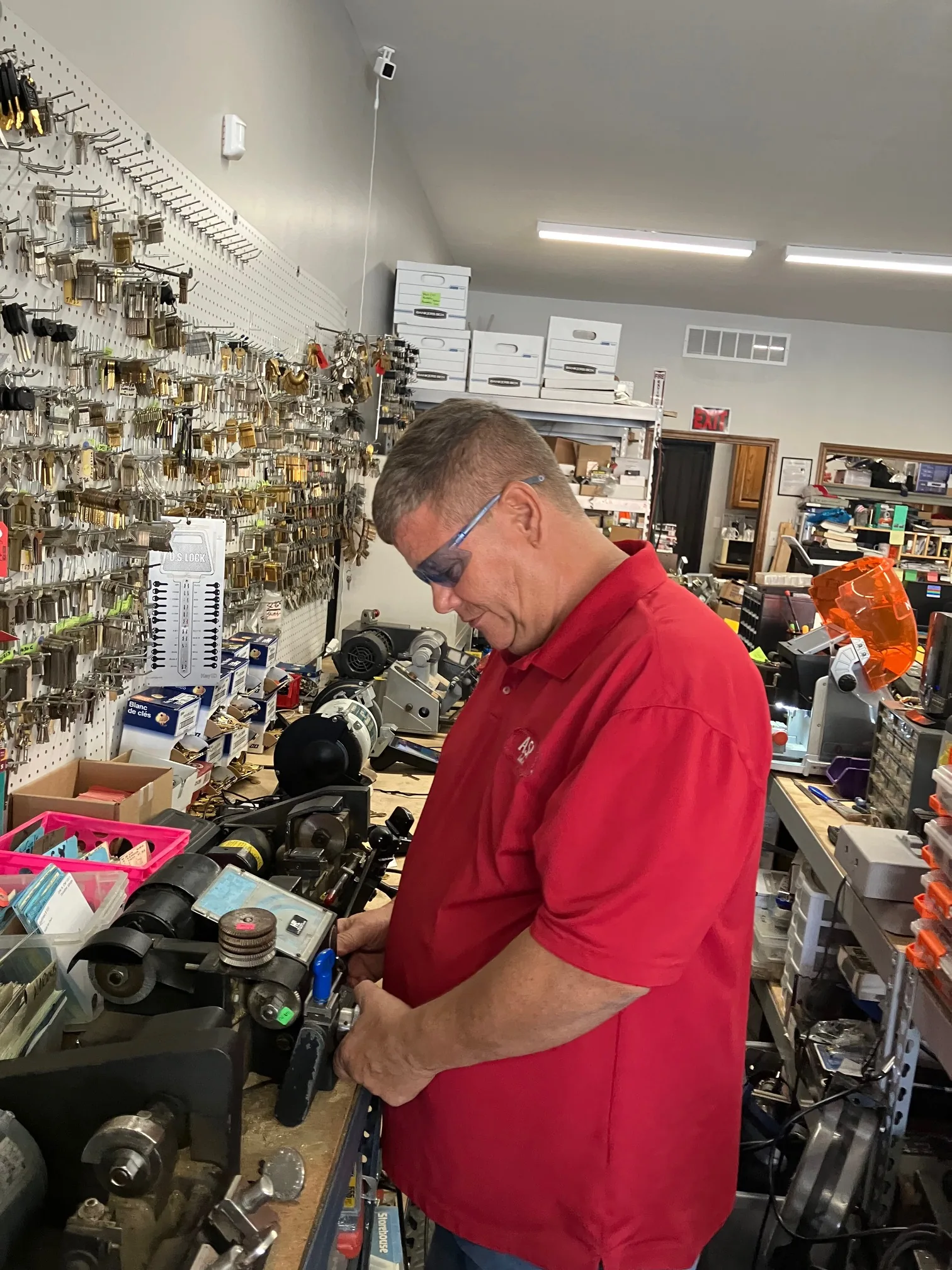 Locksmith in red shirt working on lock mechanism at workbench with pegboard of keys and tools.