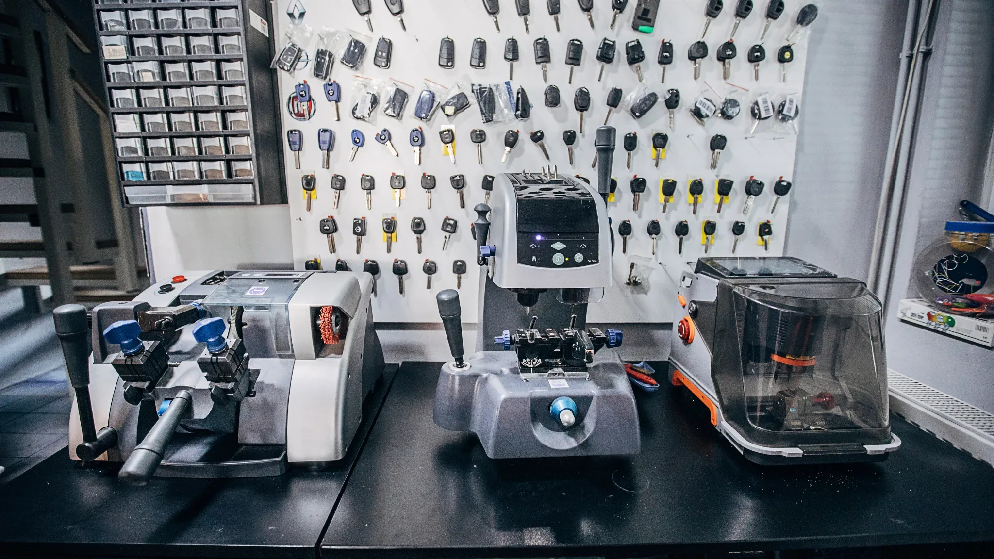 Wide view of a locksmith workbench with three key-cutting and programming machines lined up on a black counter, in front of a